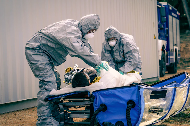 Deux personnes en combinaisons complètes de protection avec des masques sur le visage prennent en charge une personne allongée sur un brancard.