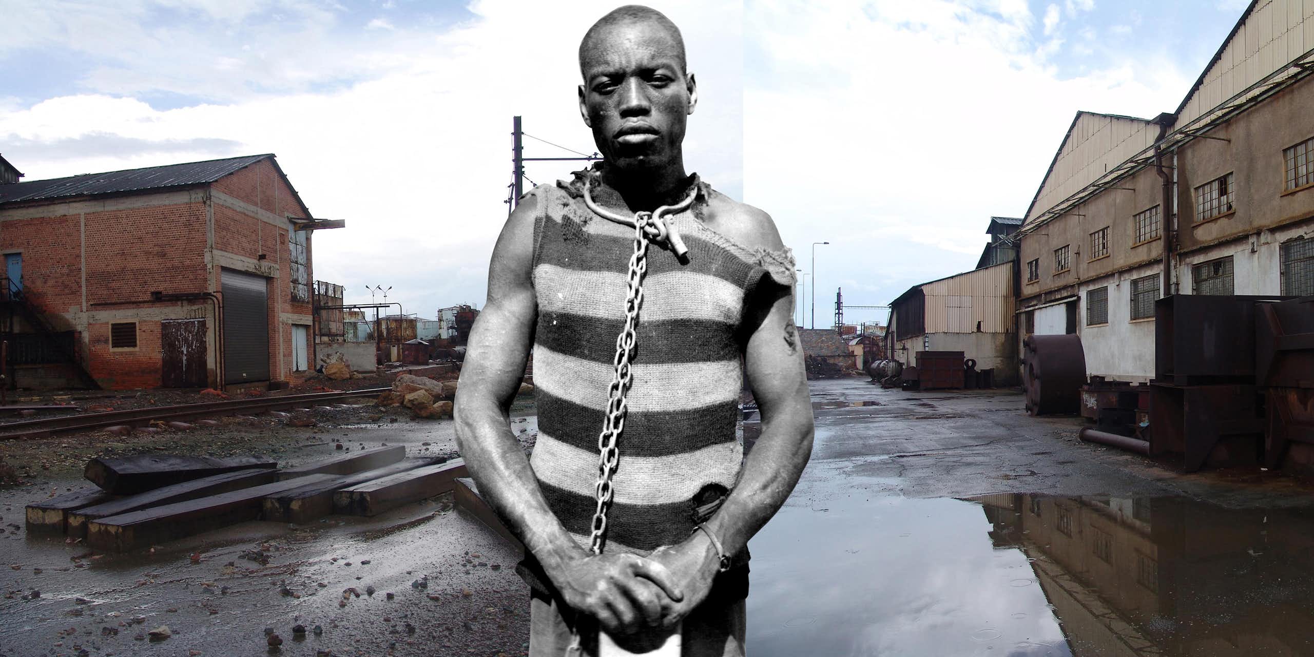 A man in a torn prison top with a shackle around his neck stands in a puddle of water, next to a buildings and railway line.