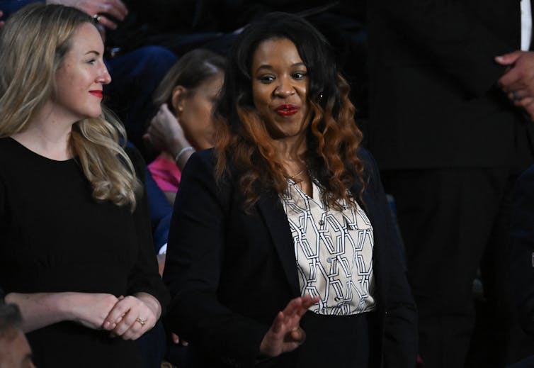 A Black woman with black hair and red highlights smiles and looks to the side, standing next to a blonde white woman, also wearing formal clothing.