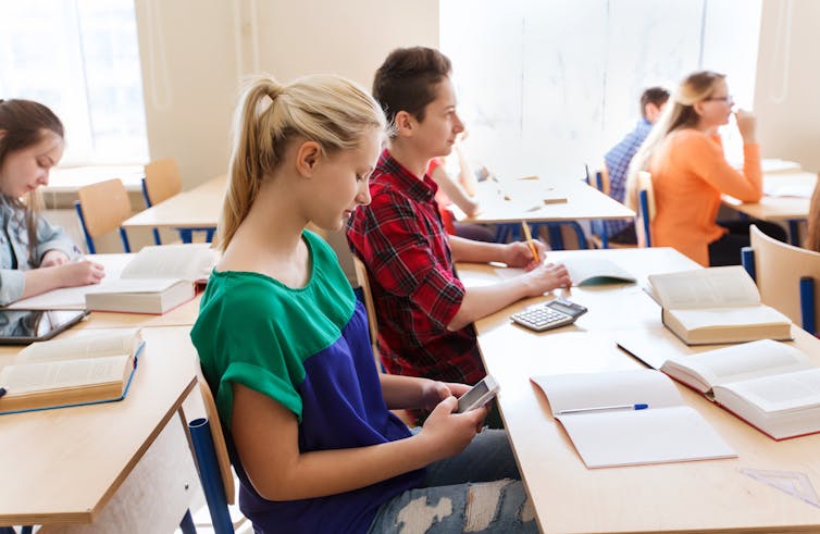 children in a classroom paying attention with the exception of one girl who is looking at her mobile phone