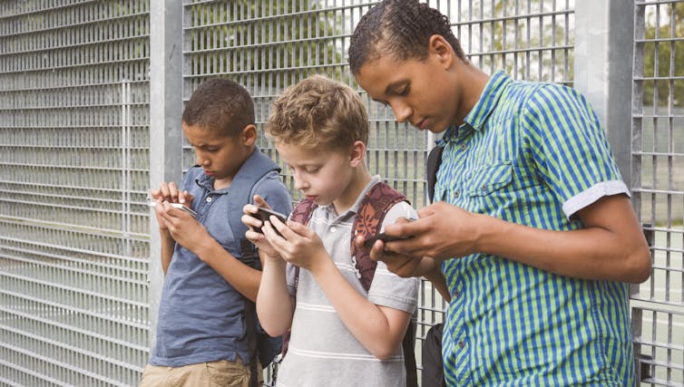three boys standing against a fence using mobile phones