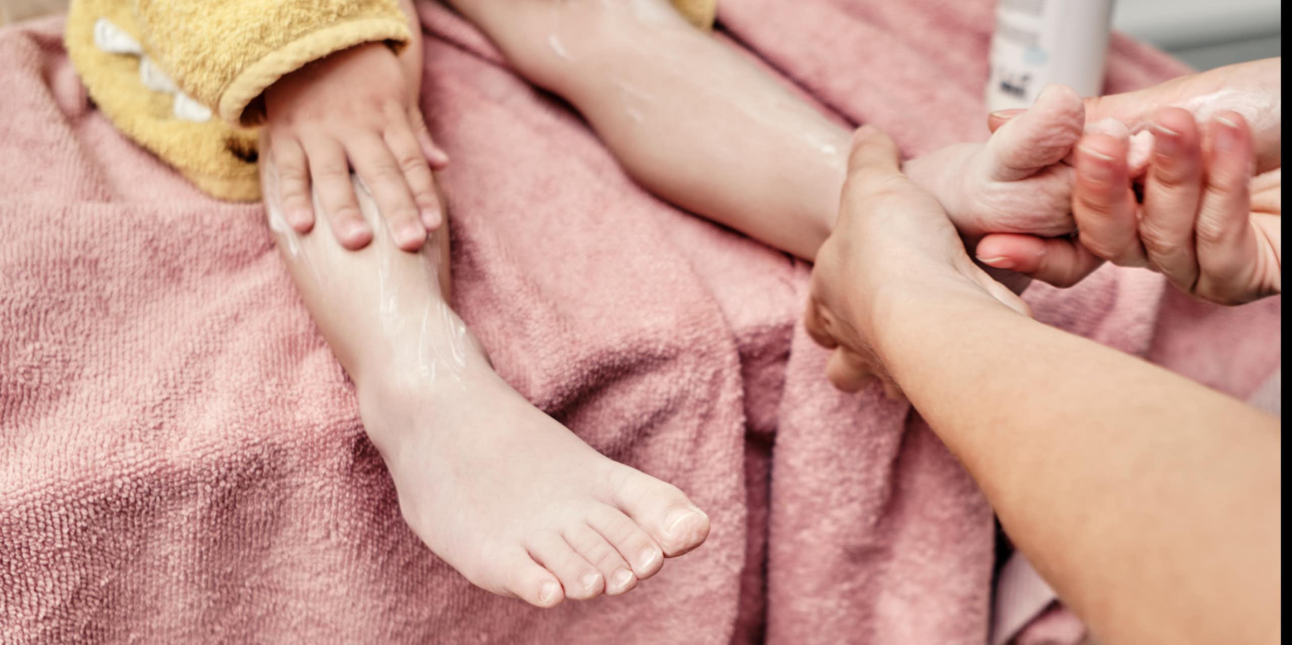 Close-up of legs of child sitting on a towel while another person slathers lotion on their foot.