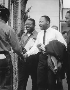 A black and white photograph of two men in collared shirts smiling outside as they shake a third man's hand.