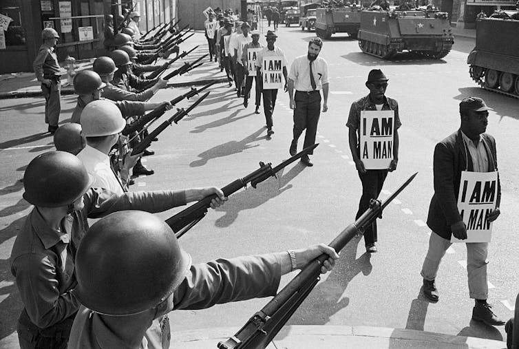 Uniformed guards lined up with rifles with bayonets on them as men walk by wearing placards that say 'I am a man.'