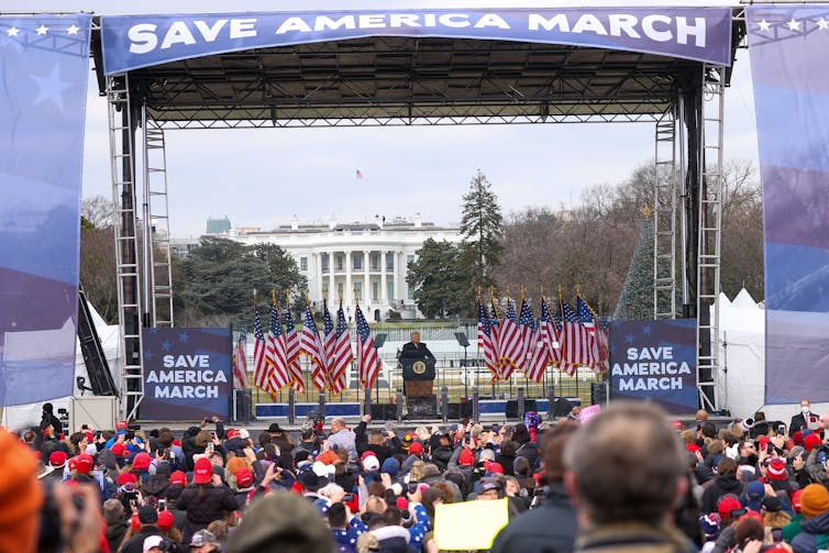 In front of a crowd stands man in a dark coat on a big stage with a banner above that says 'SAVE AMERICA MARCH.'