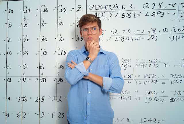 A teenage boy with a quizzical look on his face stands in front of a whiteboard filled with mathematical equations.