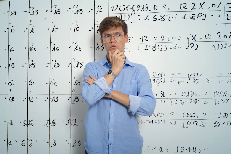 A teenage boy with a quizzical look on his face stands in front of a whiteboard filled with mathematical equations.