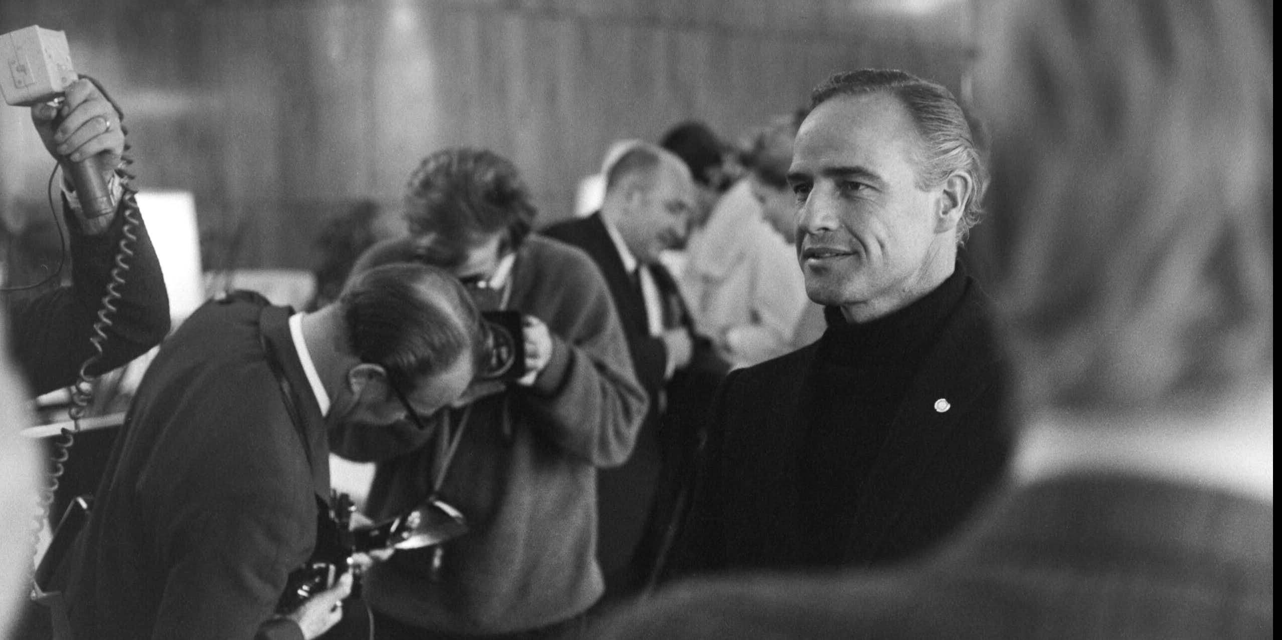 Un hombre medio sonriente rodeado de periodistas en una foto en blanco y negro.