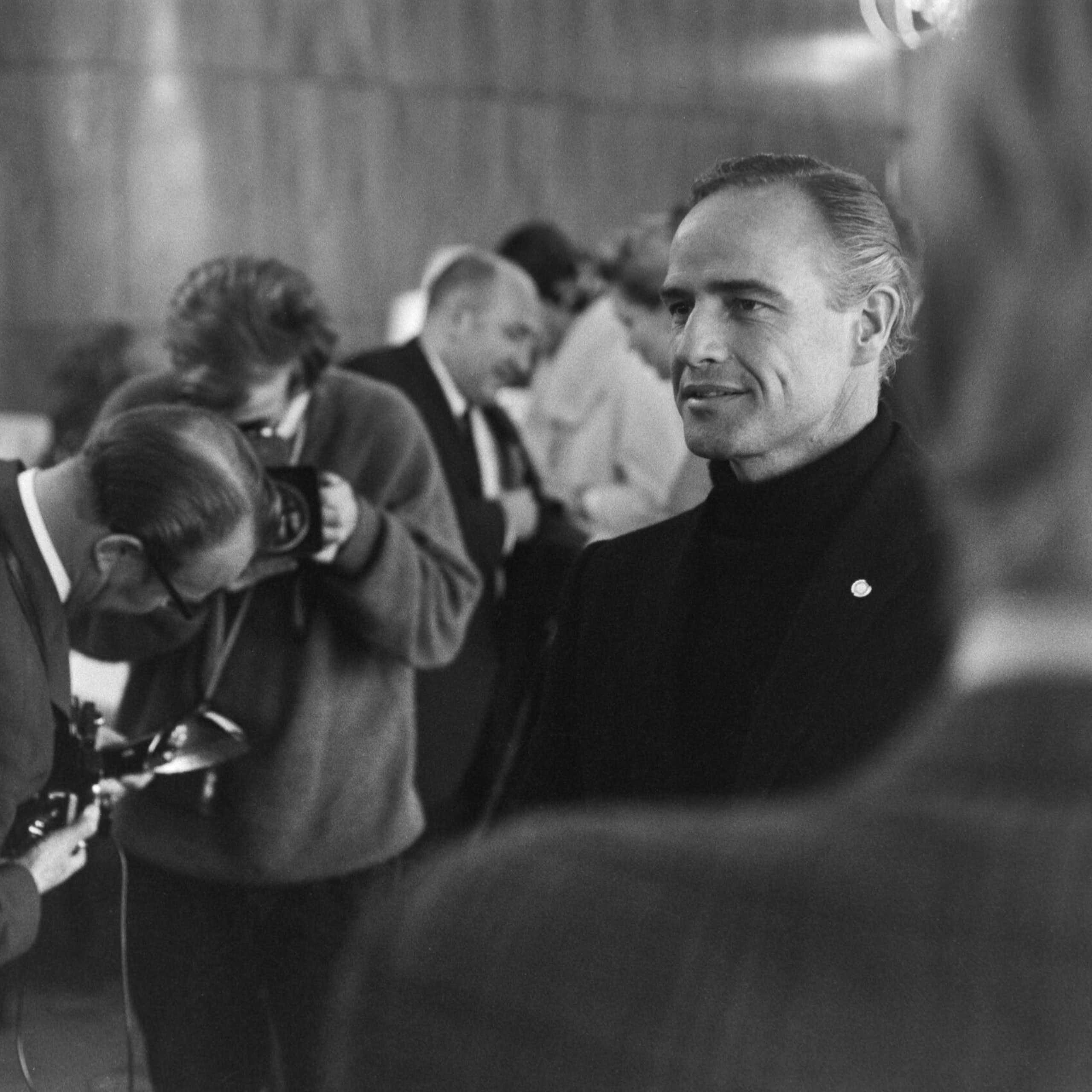 Un hombre medio sonriente rodeado de periodistas en una foto en blanco y negro.