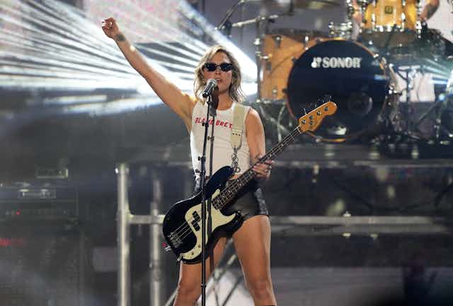 A woman on a stage in sunglasses holding a bass guitar with one arm extended in the air.