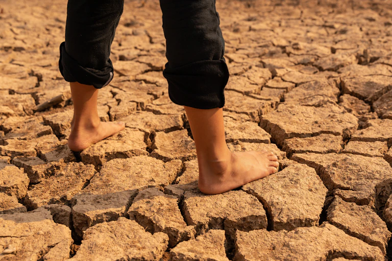 CRacked dry desert ground, young girl's legs with bare feet and black rolled up trousers standing on ground