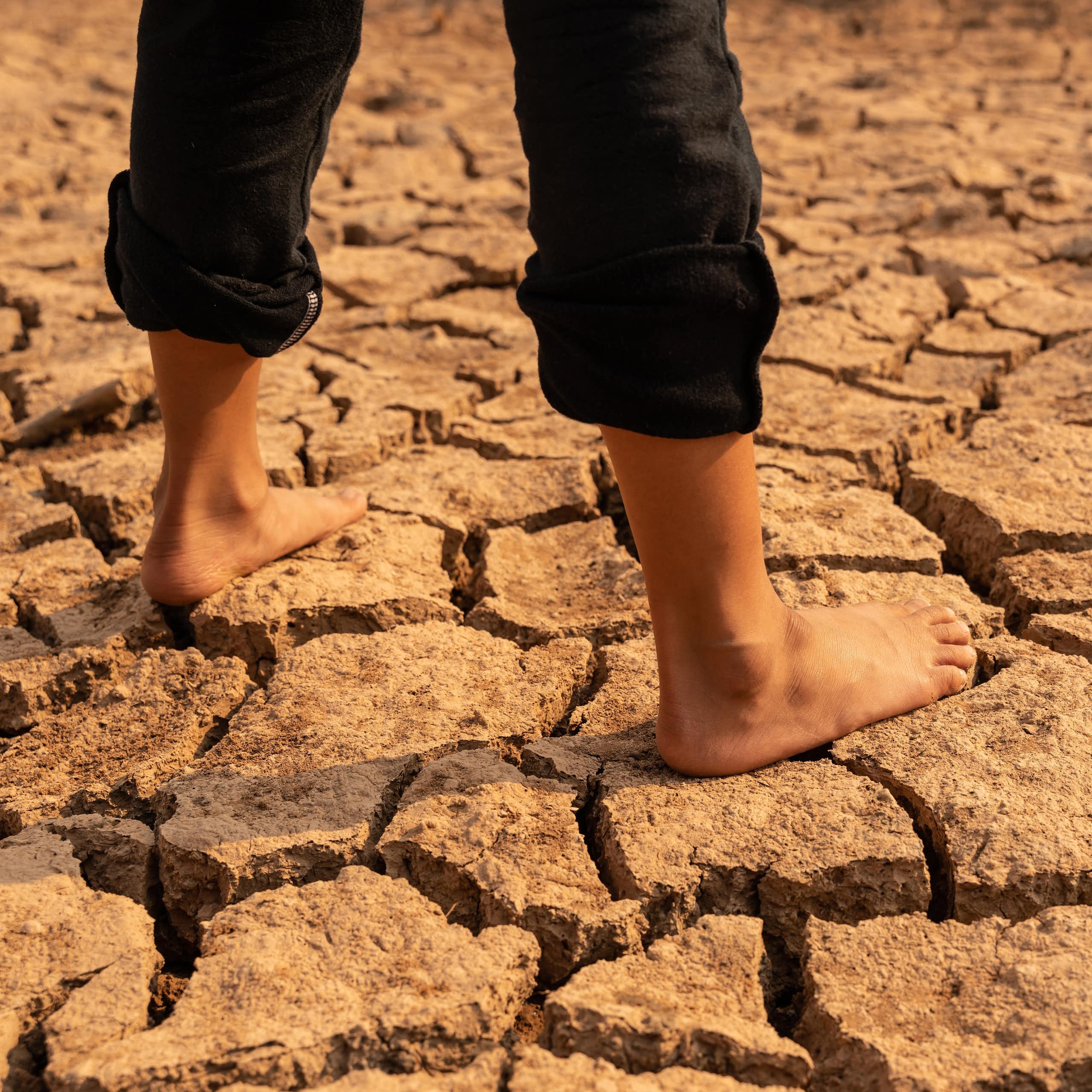 CRacked dry desert ground, young girl's legs with bare feet and black rolled up trousers standing on ground