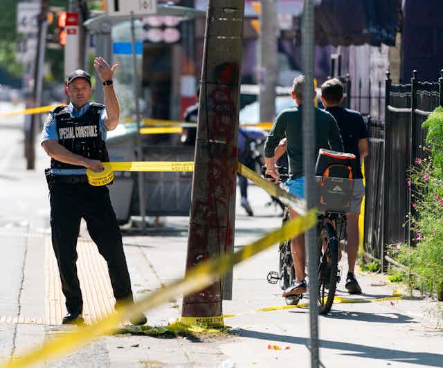 A police officer gestures with his hand as he adjusts a roll of yellow police tape on a sunny street.