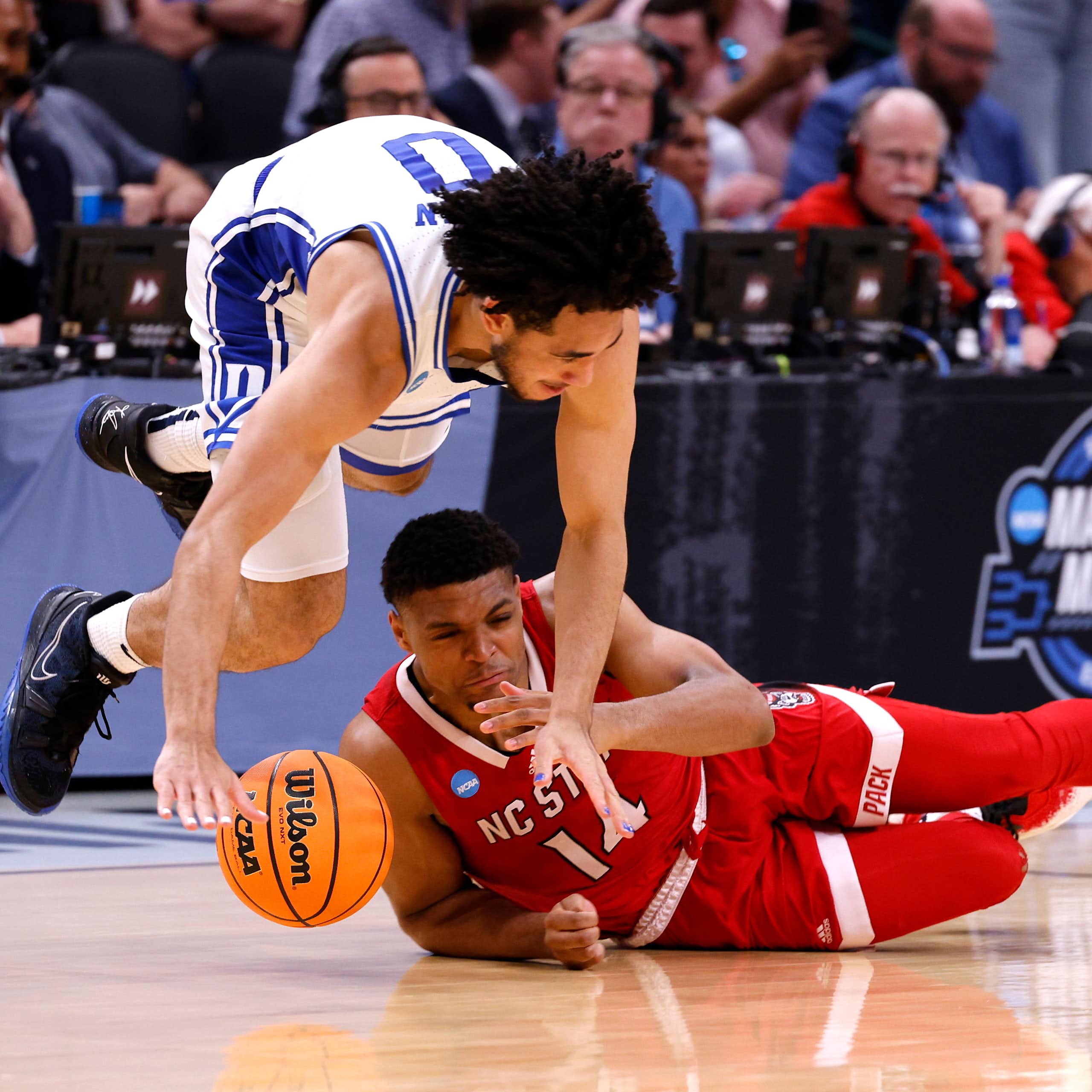 A basketball player in a white uniform with blue lettering is midair as he scrambles for a basketball with a player from the opposing team, who is wearing red, is lying on the court underneath.