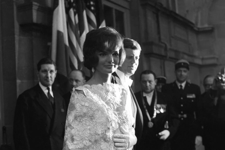 A woman dressed in a gown stands near her husband as he smiles in a spotlight.