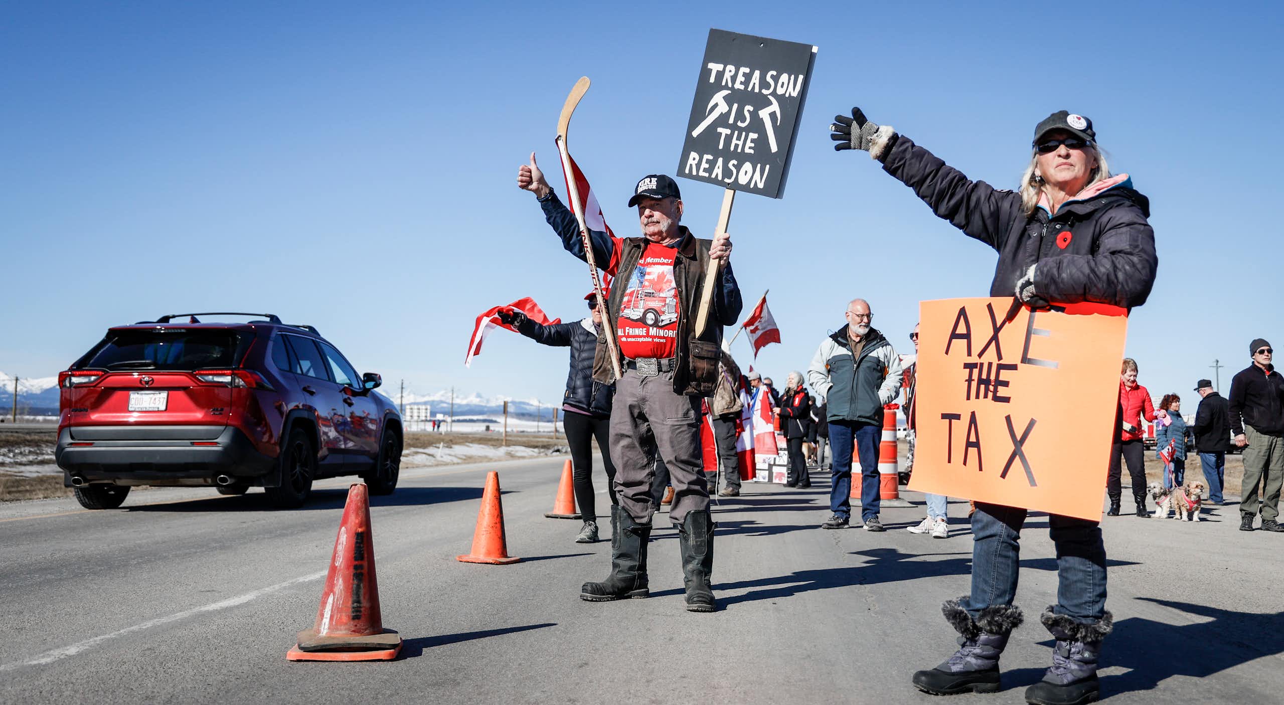 People stand at the side of a highway. One carries a sign that reads Axe the Tax.