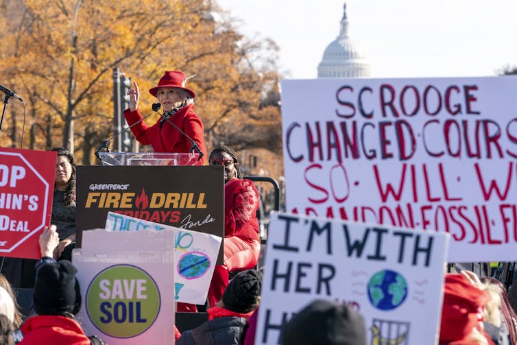 A woman wearing a red hat speaks to a crowd with signs and the U.S. Capitol in the background.