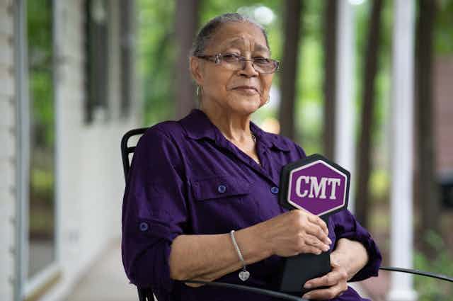 Elderly Black woman wearing glasses and a purple shirt seated in a chair.