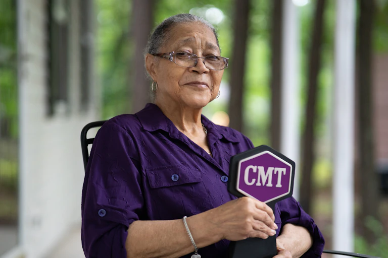 Elderly Black woman wearing glasses and a purple shirt seated in a chair.