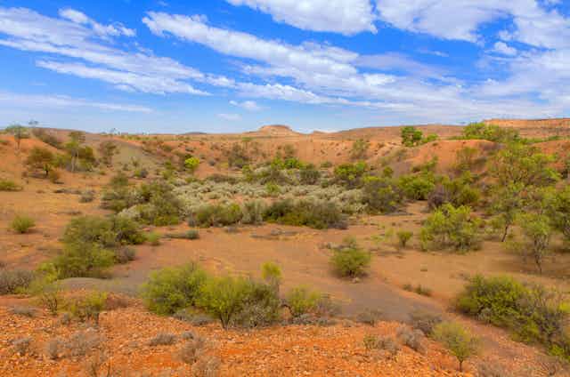 A photo of an eroded land formation in red earth with low green scrub and an odd hill in the distance, beneath a blue sky with white clouds.