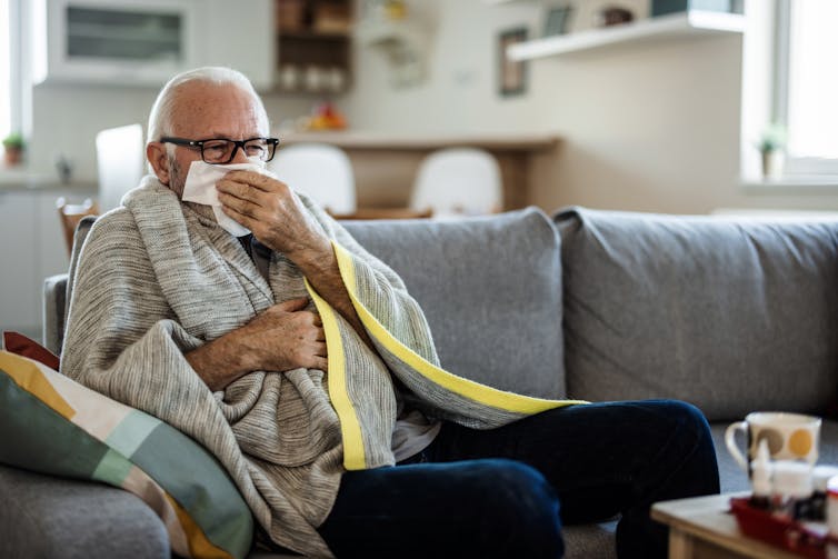 A senior man sitting on a couch using a tissue.