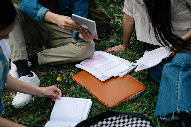 A group of young people sit on the grass with books and papers.