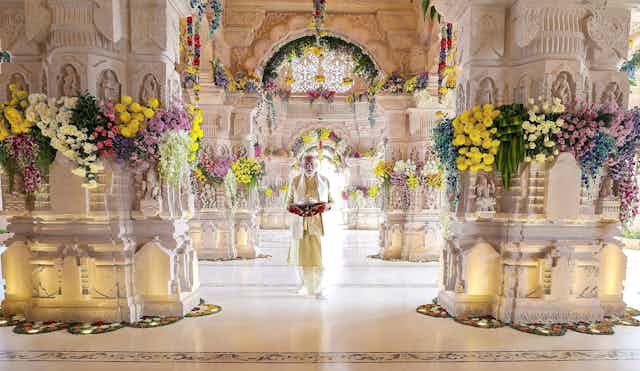 Man dressed in white suit stands inside white temple bedecked with flowers.