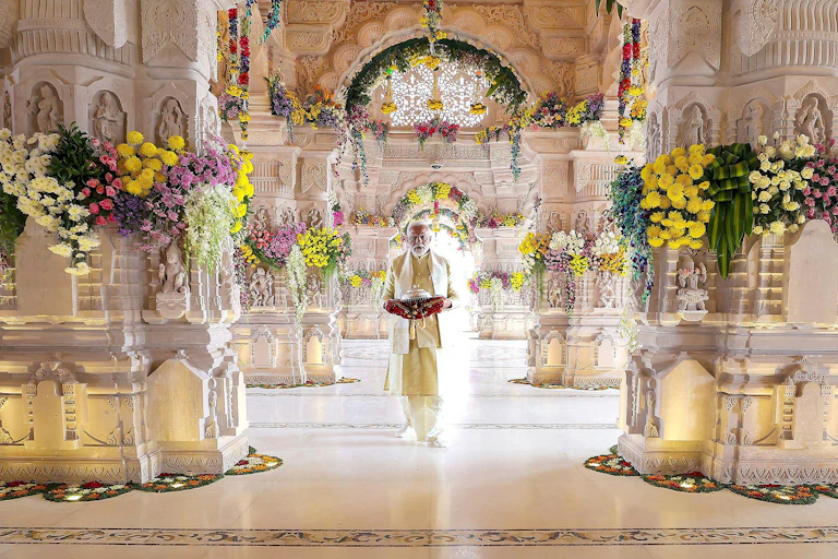 Man dressed in white suit stands inside white temple bedecked with flowers.