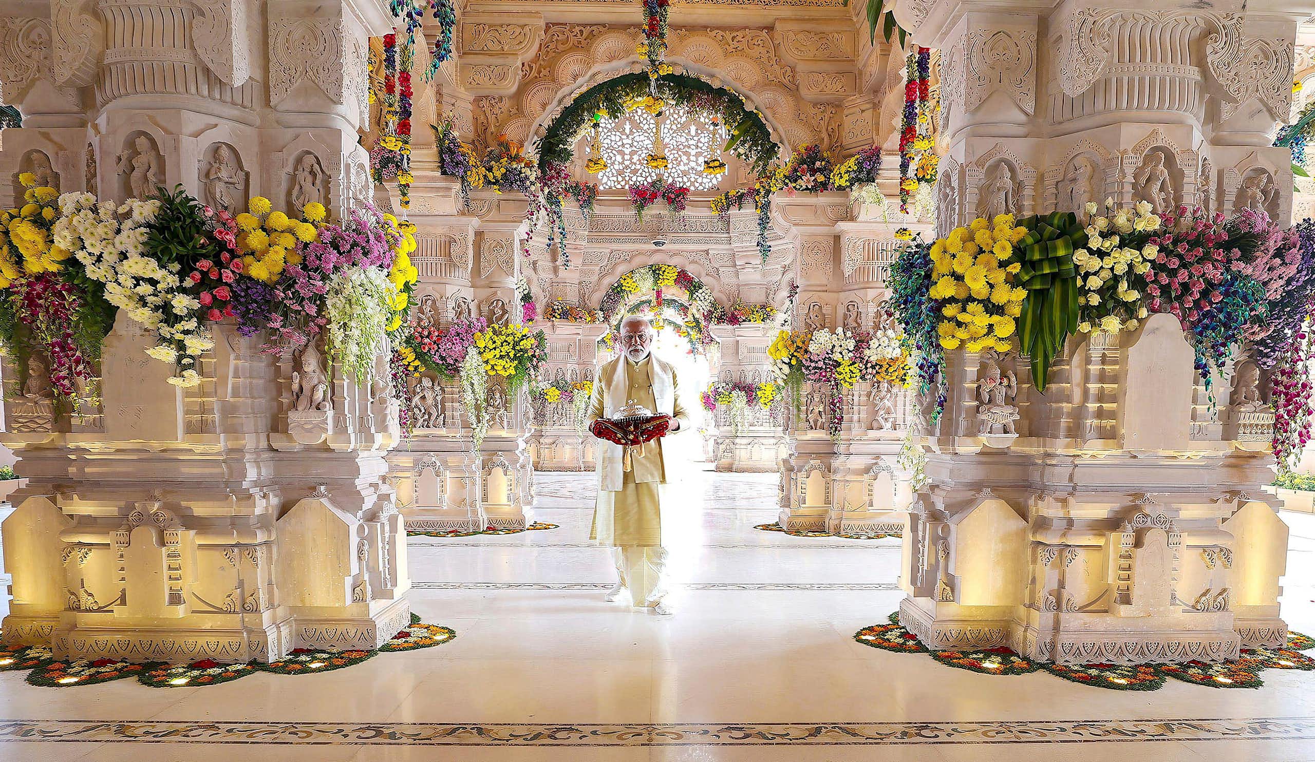 Man dressed in white suit stands inside white temple bedecked with flowers.