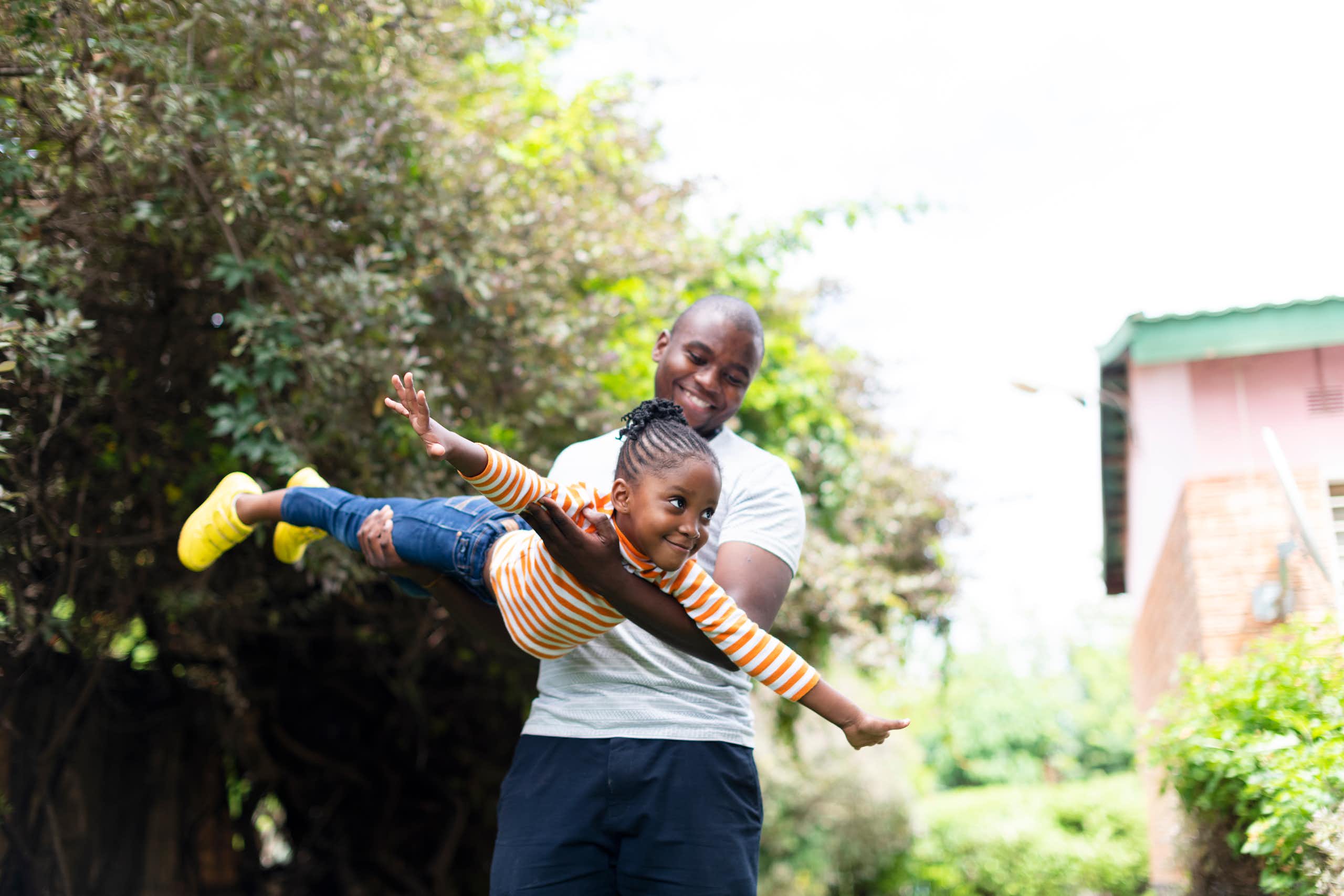 An adult man holds a young girl as she pretends she's flying. They are both smiling broadly