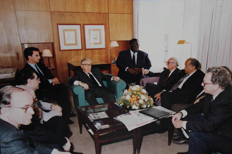 A group of nine men seated in a room with one man at the centre of the room in a U-shape