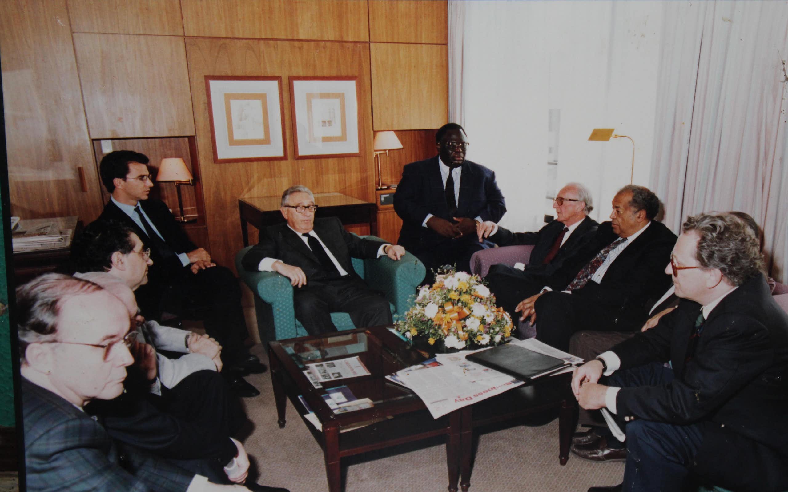 A group of nine men seated in a room with one man at the centre of the room in a U-shape