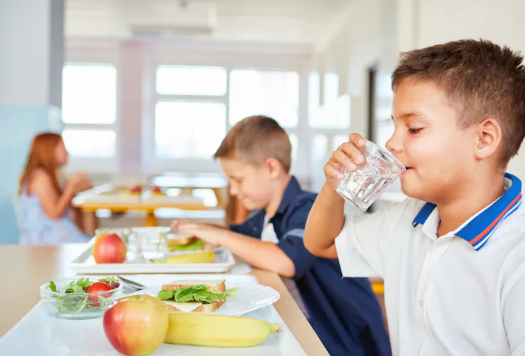 A boy drinks a glass of water in a school cafeteria with other children nearby. Fruit is on the tray in front of him.