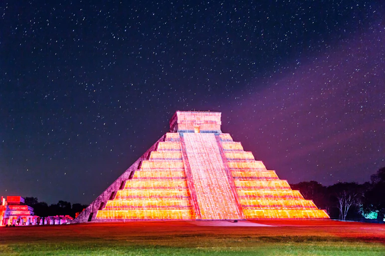 A stone pyramid with many tiers, lit up in shades of yellow and pink under a night sky with lots of stars.