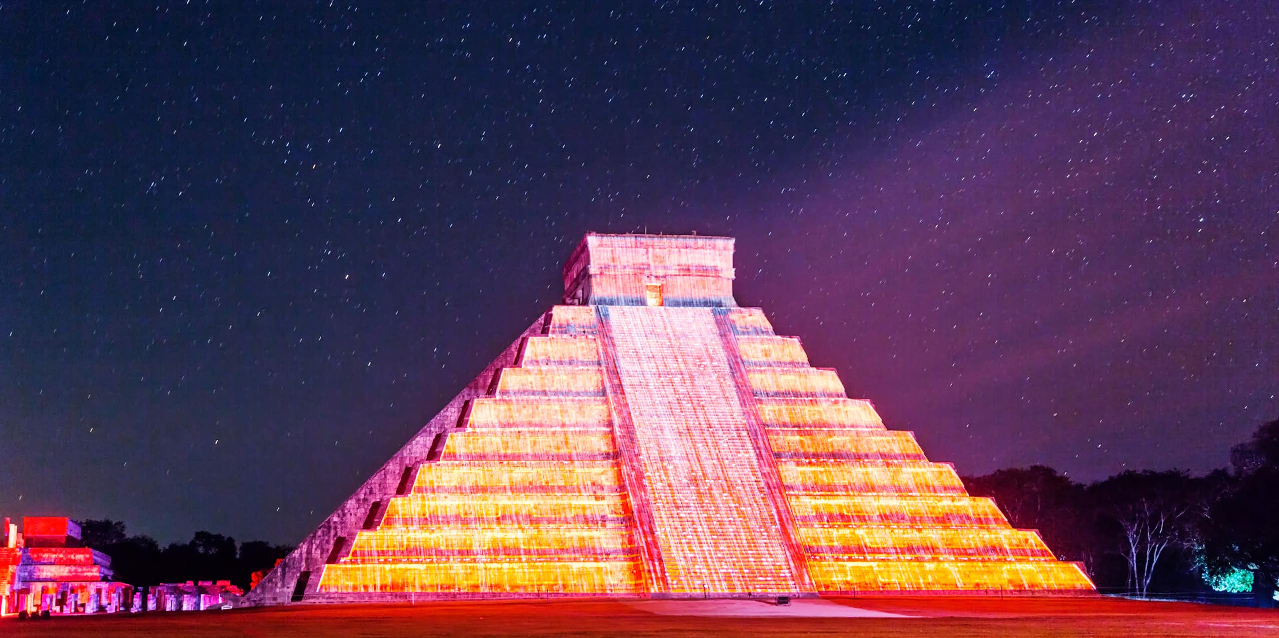 A stone pyramid with many tiers, lit up in shades of yellow and pink under a night sky with lots of stars.
