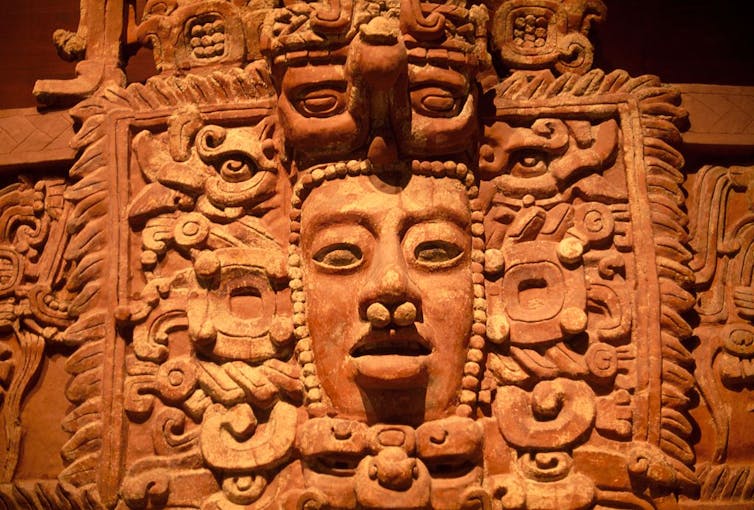 A large clay-colored decoration with intricate carvings and a face with eyes, a pierced nose and a mouth.