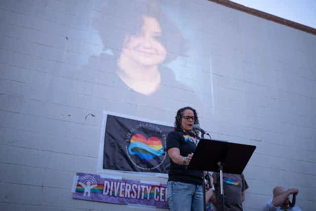 A woman with dark hair and a black shirt with a rainbow on it stands at a podium in front of a brick wall with a photo of a smiling teenager. 