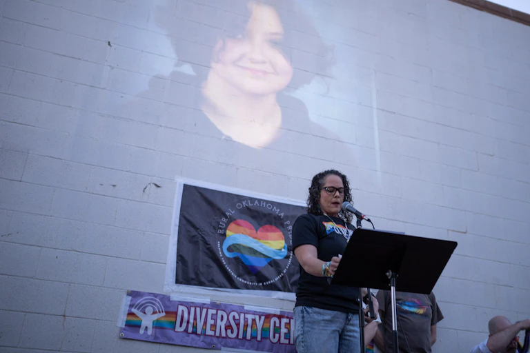 A woman with dark hair and a black shirt with a rainbow on it stands at a podium in front of a brick wall with a photo of a smiling teenager.