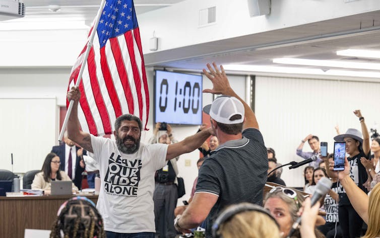 A man waves a large American flag and wears a white shirt that says 'Leave our kids alone.' He stands in a crowded room