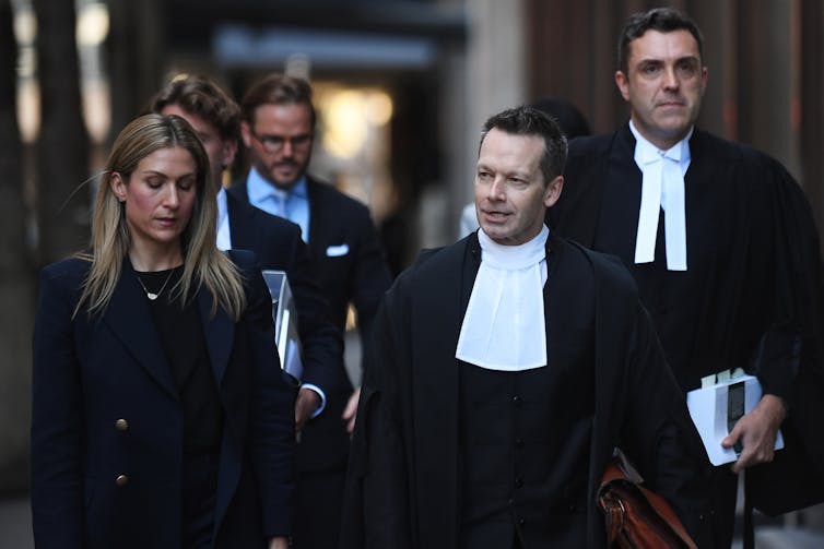 Two male lawyers in robes walk down the street accompanied by a woman in black