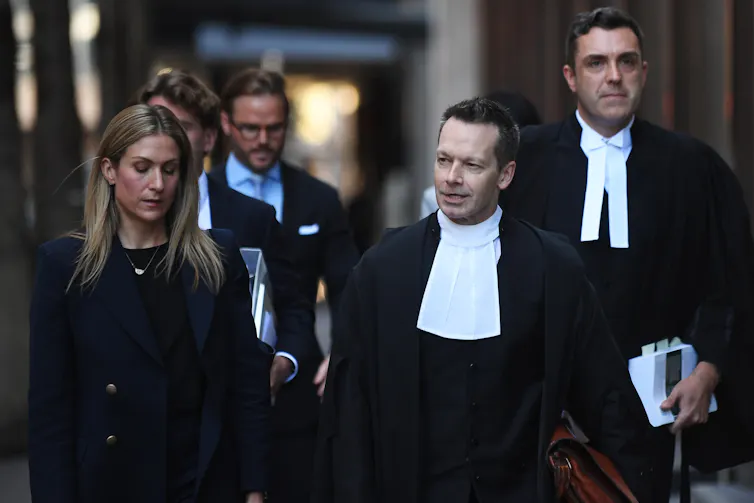 Two male lawyers in robes walk down the street accompanied by a woman in black