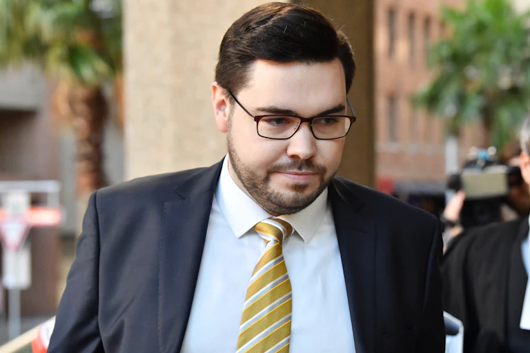 A dark-haired man with glasses in a suit and tie enters a courtroom