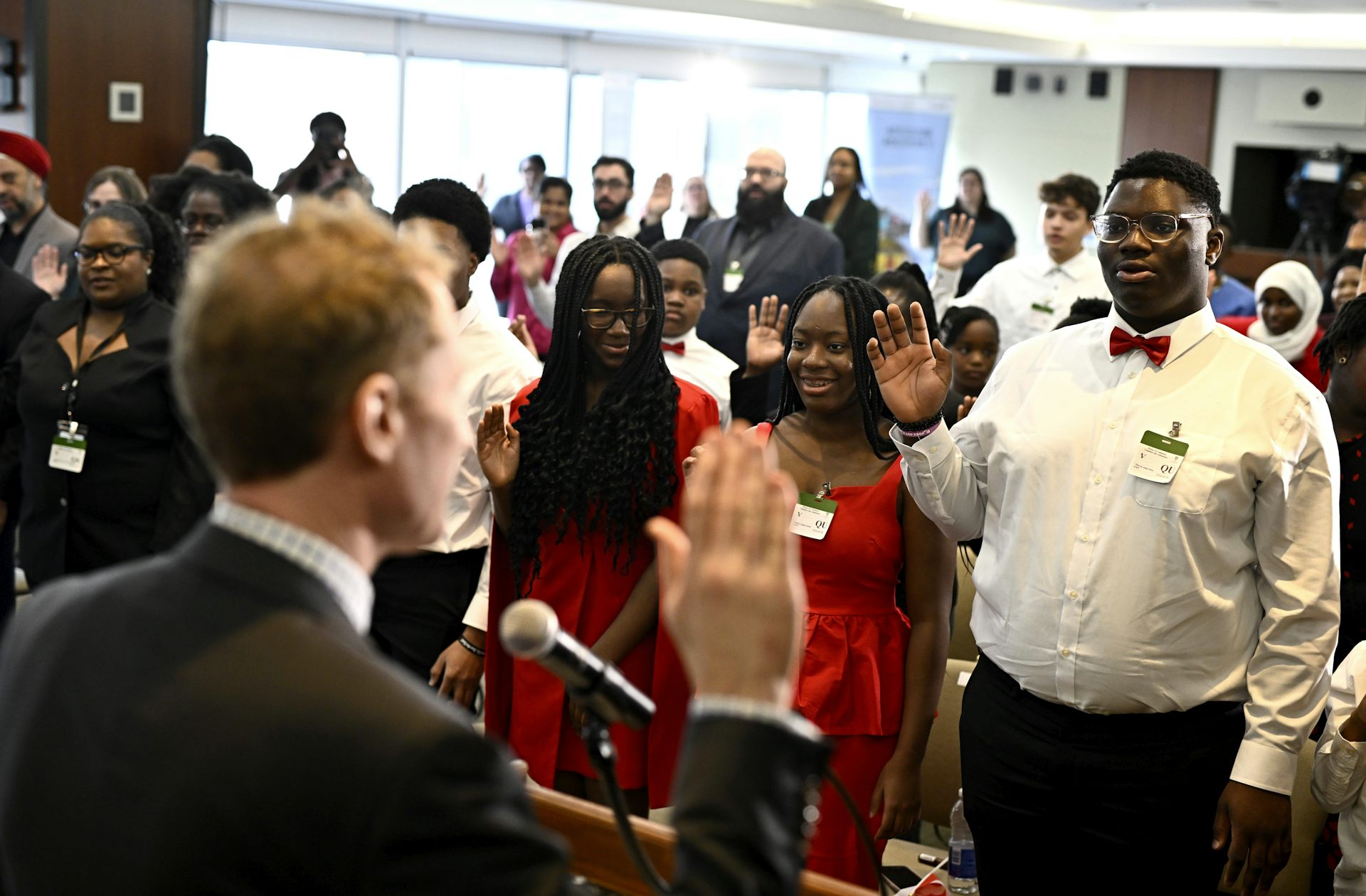 A group of people wearing formal attire raise their right hands while looking at a man who has his back to the camera