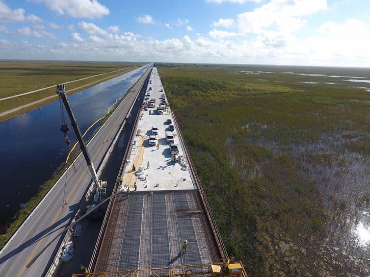 An elevated roadway under construction over a far-reaching marsh area.