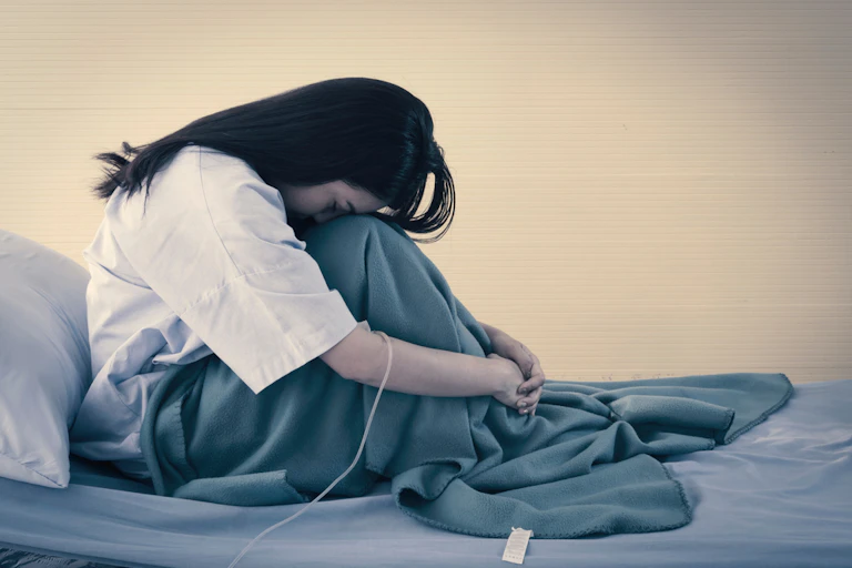 Young woman sitting on hospital bed, head on knees, IV drip in