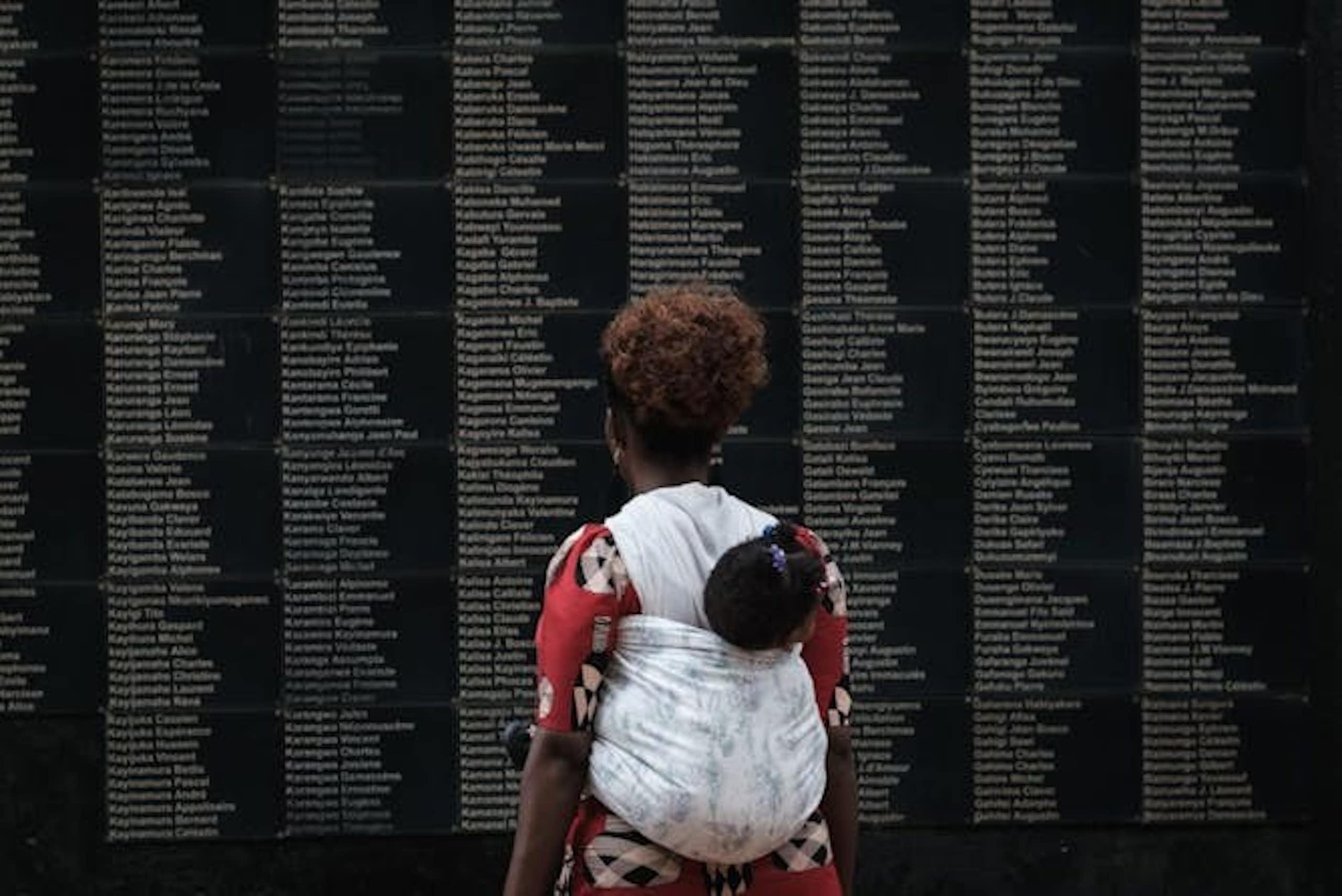 Une femme debout devant un mur noir sur lequel figure un texte blanc, un bébé attaché dans le dos.
