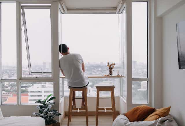 View from behind a young man sitting on a stool in a modern city flat, looking out of a large window at the cityscape. He is leaning his shoulder and head against the wall.