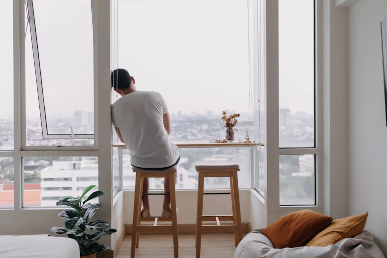 View from behind a young man sitting on a stool in a modern city flat, looking out of a large window at the cityscape. He is leaning his shoulder and head against the wall.