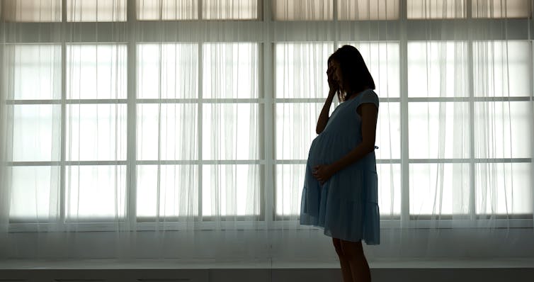 A silhouette of a young pregnant woman in a dress standing in front of a wall of bright, curtain-covered windows with her hand over her face