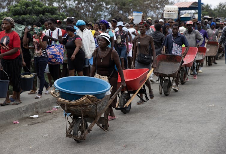 A queue of people walking down a road with empty buckets and wheelbarrows.